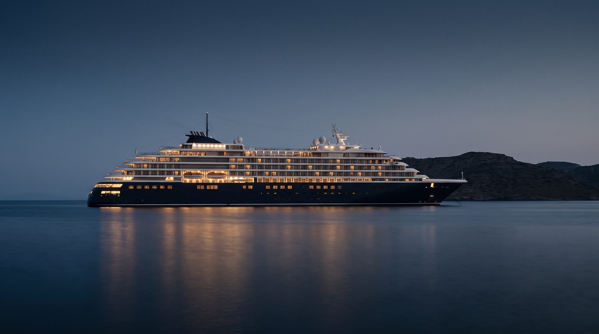 A large luxury yacht lying at anchor at blue hour off a Greek island, its suites lit from within.